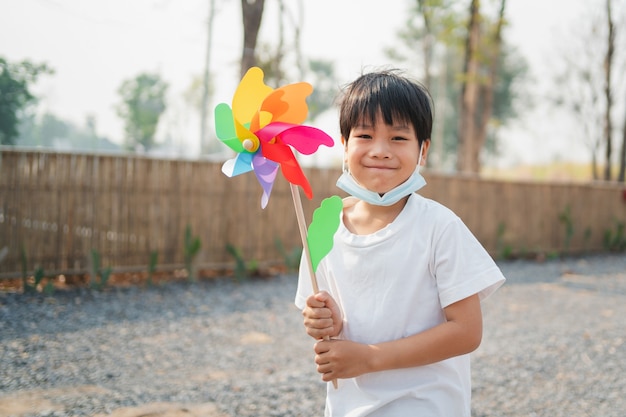Un garçon tenant un jouet moulin à vent marchant joyeusement dans le parc et portant un masque pour se protéger contre le virus