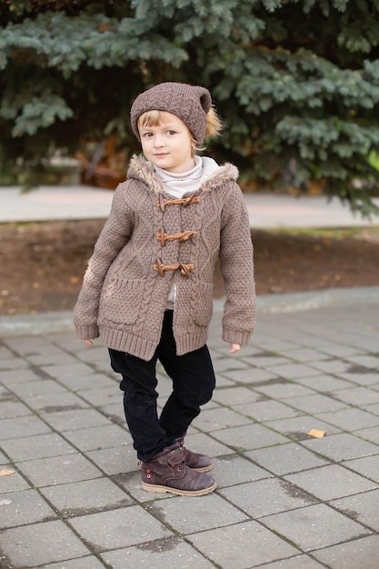 un garçon en tricot, un chapeau et une veste marron se tient dans la rue