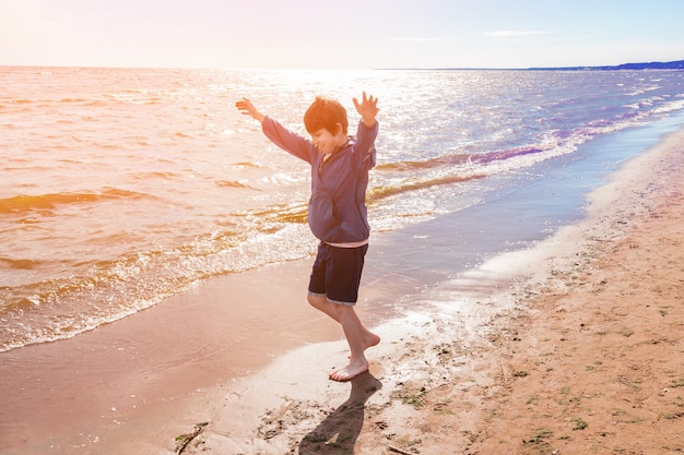 Garçon en veste bleue et short aux pieds nus courant joyeusement avec les mains sur le sable au bord de la mer, journée ensoleillée