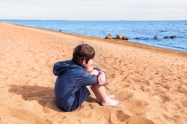 Garçon en veste bleue et short pieds nus assis sur le sable au bord de la mer, journée ensoleillée.
