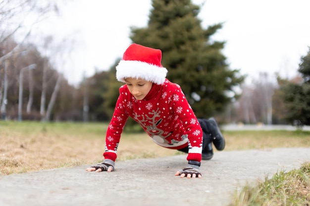 Un garçon vêtu d'un pull de Noël chaud avec un cerf du Nouvel An et un bonnet de Noel s'apprêtait à effectuer un exercice de pompes dans un parc de la ville sur les pistes