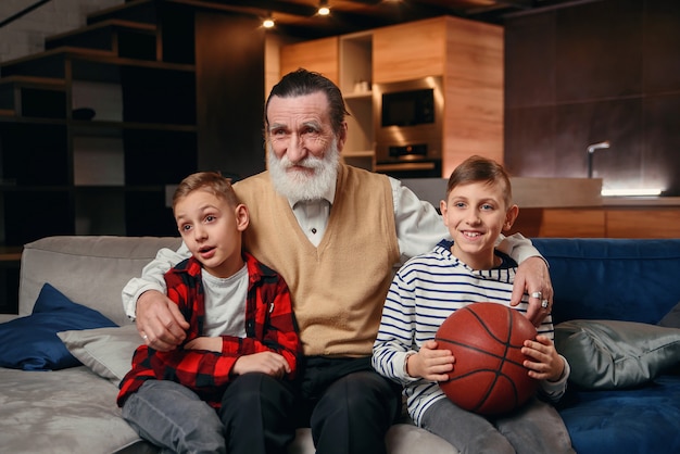 Garçons sur canapé avec leur grand-père applaudir pour un match de basket et tenant un ballon de basket. D'énormes fans de sport.