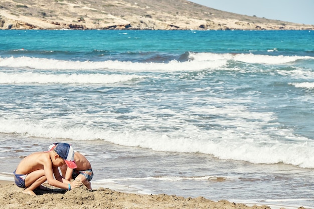 Les garçons construisent un château de sable sur la ligne de surf contre les vagues de la mer d'azur roulant avec de la mousse épaisse sur Prasonisi
