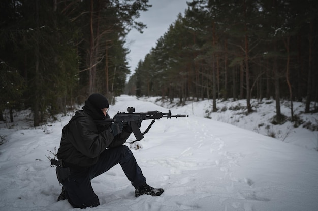 Photo un garde frontière assis sur son genou vise avec un fusil d'assaut ak-12 dans la forêt