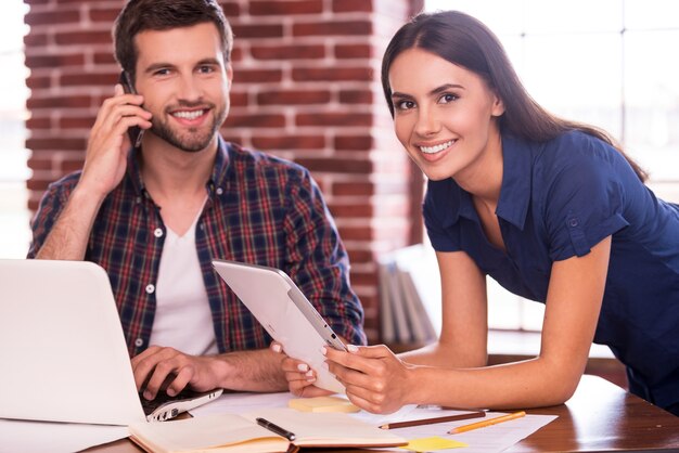 Garder une atmosphère amicale au travail. Joyeux jeune homme et femme assis sur le lieu de travail et souriant tandis qu'une femme tenant une tablette numérique et un homme parlant au téléphone portable