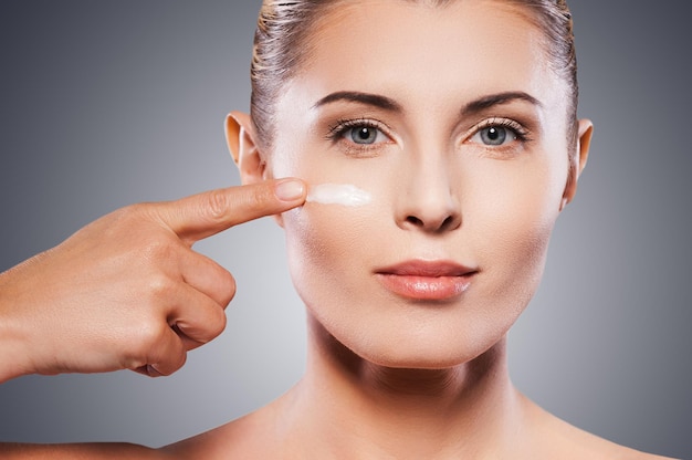 Garder sa peau jeune et lisse. Portrait d'une femme mûre torse nu répandant de la crème sur le visage et souriant en se tenant debout sur fond gris