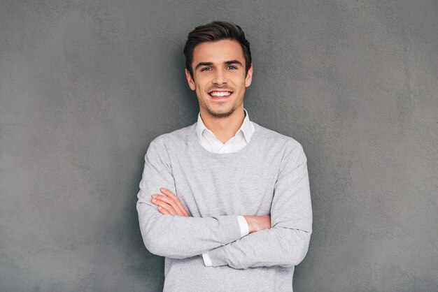 Garder le sourire. Jeune homme confiant gardant les bras croisés avec le sourire en se tenant debout