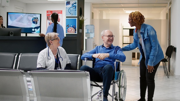 Gardien aidant un homme âgé handicapé lors d'une visite de contrôle à la réception de l'hôpital. Femme qui allaite une patiente handicapée en fauteuil roulant pour prendre rendez-vous pour une consultation médicale.