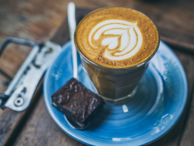 Garniture de café Piccolo Latte avec art floral de lait dans un petit verre avec un morceau de gâteau brownies fait maison sur une plaque en céramique bleue sur une table en bois