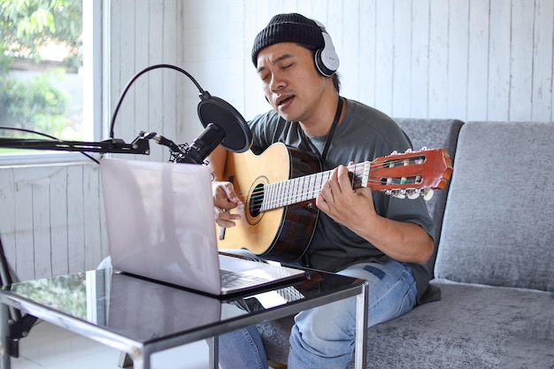 Un gars asiatique jouant de la guitare enregistrant un podcast audio en studio à l'aide d'un microphone et d'un ordinateur portable. Une personne