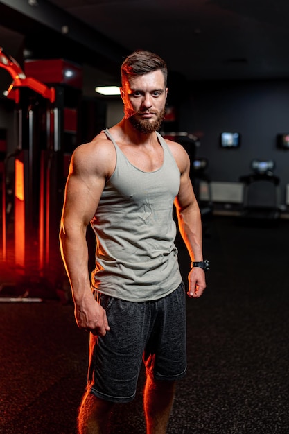 Le gars athlétique regarde la caméra. Gros muscles, abdominaux forts. Beau modèle sportif sur le fond de la salle de gym.