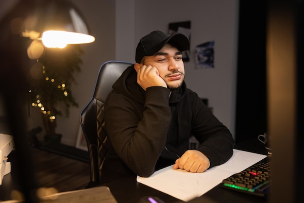 Un gars attentionné en casquette et sweat-shirt est assis devant un ordinateur qui travaille à distance de chez lui la nuit