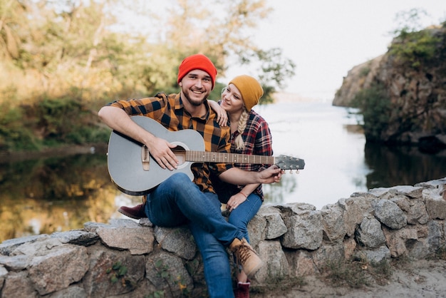 Un gars au chapeau brillant joue de la guitare avec une fille sur fond de rochers de granit et d'une rivière