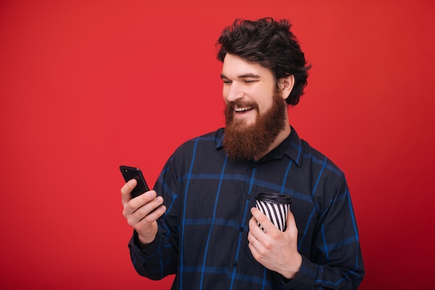 Un gars barbu à l'aide d'un smartphone tout en tenant une tasse de café, sur le mur rouge