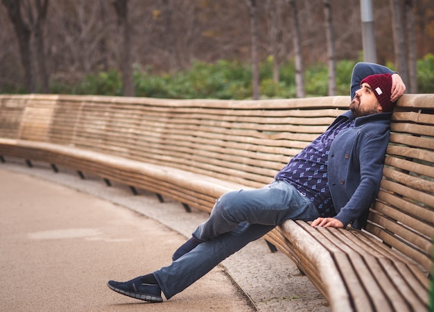 Gars barbu dans un chapeau de laine et un pull assis sur un banc de parc de détente