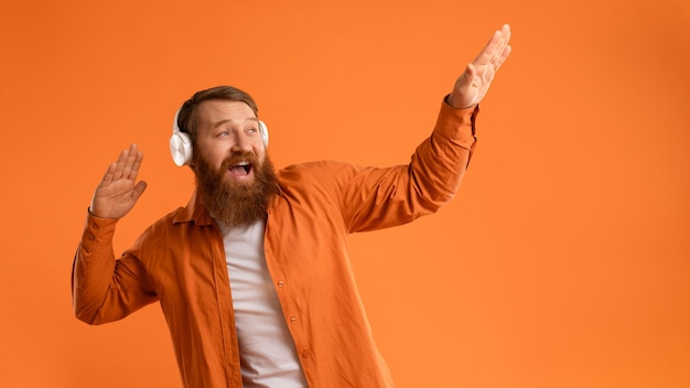 Un gars barbu positif avec un casque sans fil dansant sur un mur orange.