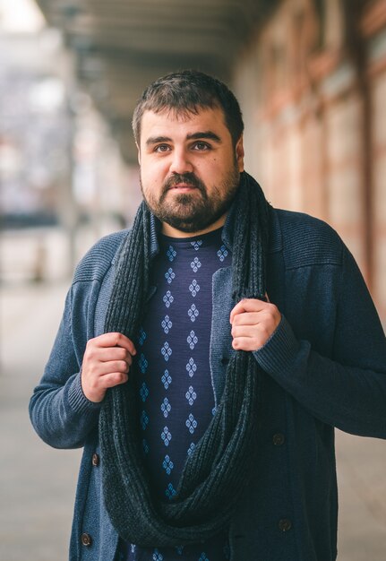 Gars barbu en pull bleu debout dans la rue avec ses mains sur son écharpe en regardant la caméra