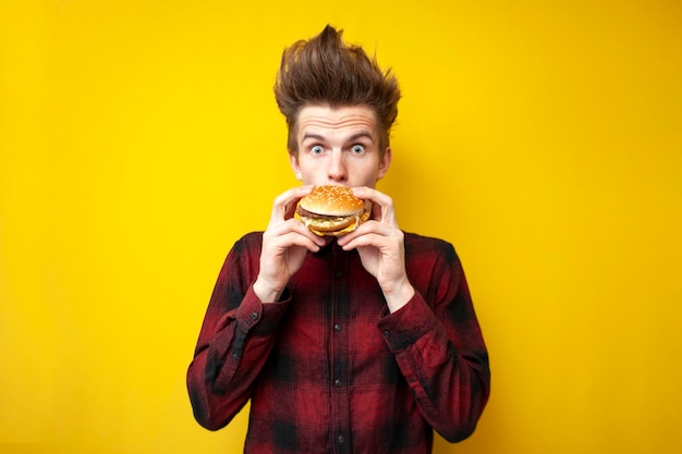 Un gars choqué en train de manger un hamburger sur un fond isolé jaune a surpris un hipster en train de manger de la restauration rapide savoureuse