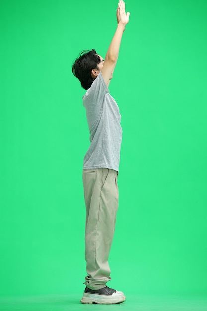 Un gars dans un T-shirt gris sur un fond vert à pleine hauteur a levé les mains