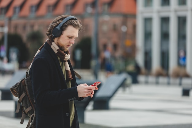 Un gars élégant en écharpe et manteau écoute de la musique sur le service de streaming en utilisant un téléphone portable dans la rue de Wroclaw Pologne