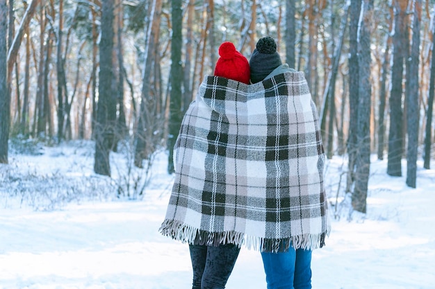 Un gars et une fille en chapeaux tricotés avec des bubons debout en hiver dans le parc sont recouverts d'une couverture chaude. Vue arrière.