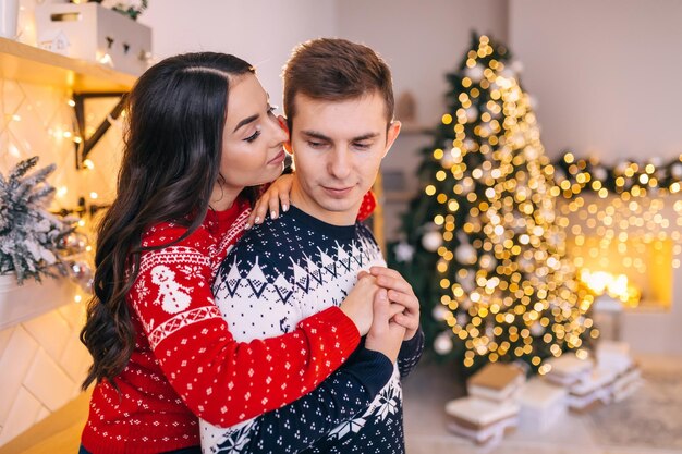 Un gars avec une fille dans un pull et un jean s'embrasse et s'embrasse sur le fond d'un sapin de Noël dans la chambre