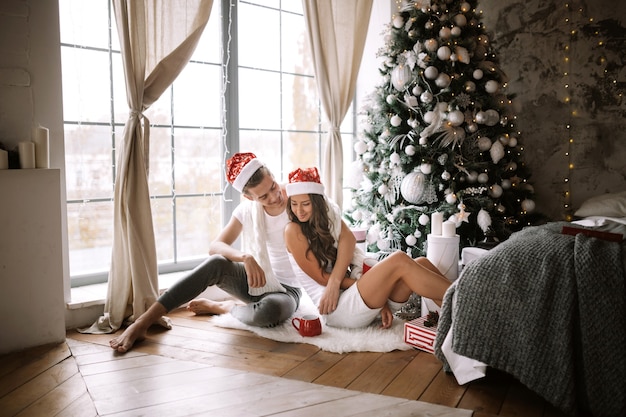 Un gars et une fille heureux en t-shirts blancs et chapeaux de père Noël sont assis et s'embrassent dans la pièce par terre devant la fenêtre à côté de l'arbre du Nouvel An, des cadeaux et des bougies.