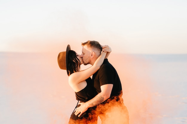 Un gars et une fille en vêtements noirs s'embrassent et courent sur le sable blanc avec de la fumée orange