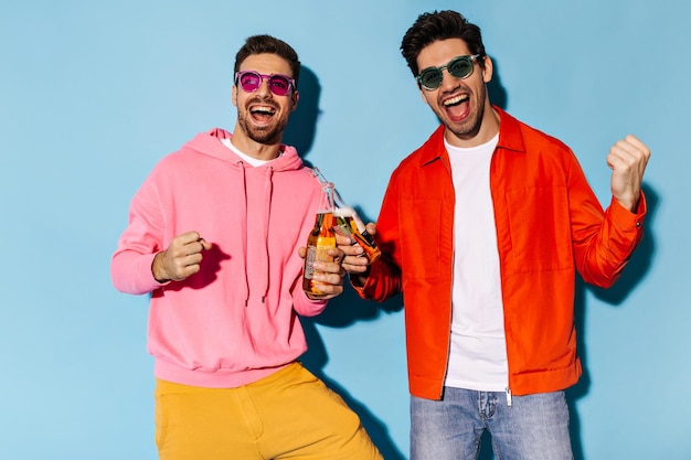 Des gars gais barbus excités en veste orange et sweat à capuche rose se réjouissent de s'amuser et de tinter des bouteilles de bière sur fond bleu