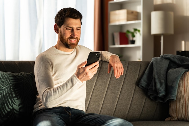 Un gars insouciant en vêtements de maison est assis sur un canapé confortable. Il passe du temps avec son téléphone portable dans son appartement. Stock photo