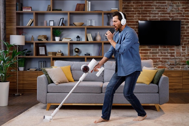 Un gars joyeux l'homme nettoie l'appartement Passer l'aspirateur avec des écouteurs en dansant et en chantant dans un smartphone au lieu d'un microphone Le nettoyage de la maison est facile