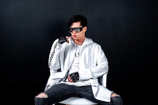 un gars à lunettes noires est assis sur une chaise sur un fond sombre de béton dans un studio; dans le studio est un jeune homme à lunettes noires