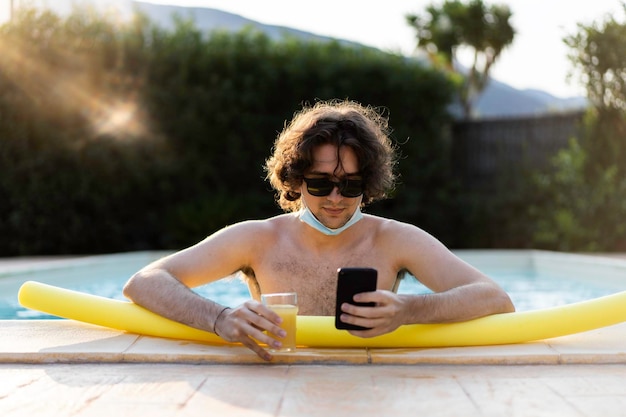 Un gars avec un masque médical s'amusant au bord de la piscine en tapant avec un smartphone et en buvant du jus d'orange pendant l'été 2021 après l'épidémie de coronavirus Un gars profitant de la vie seul en raison des restrictions estivales