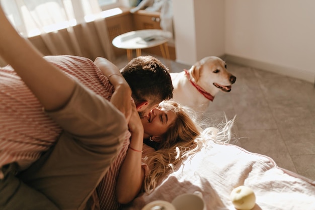 Le gars de Nrunet et sa fille bouclée sont doux et tendrement embrassés allongés sur le lit dans leur appartement pendant que leur Labrador est assis à proximité