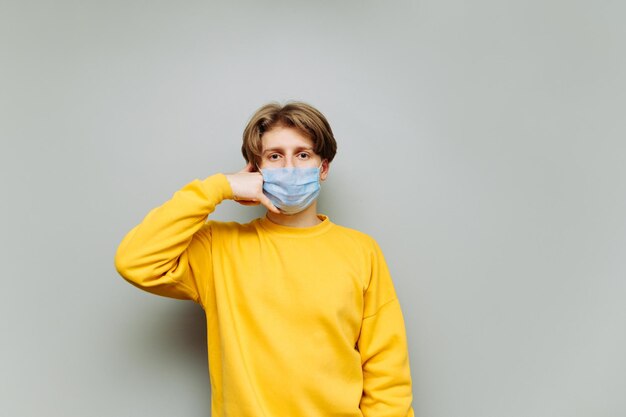 Un gars portant un masque médical sur son visage en sweat-shirt jaune montre un geste d'appel et regarde la caméra