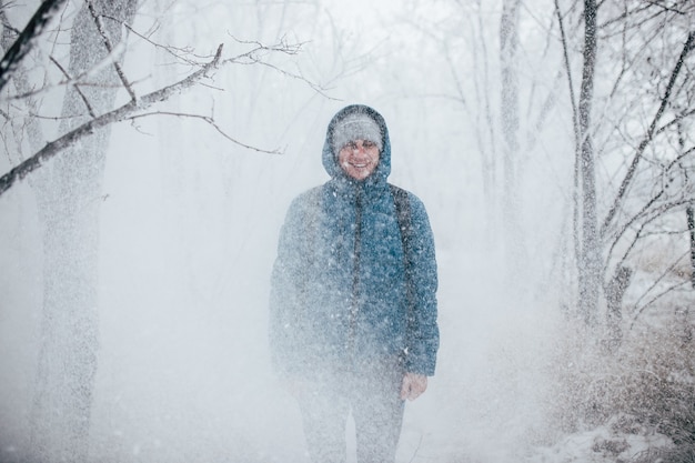 Un gars portant une veste à capuche se tient dans une forêt enneigée, la neige tombe sur lui d'en haut.