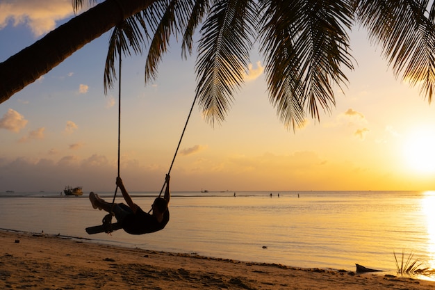 Le gars profite du coucher de soleil sur une balançoire sur la plage ptropicale. Silhouettes d'un gars sur une balançoire accrochée à un palmier, regardant le coucher du soleil dans l'eau.