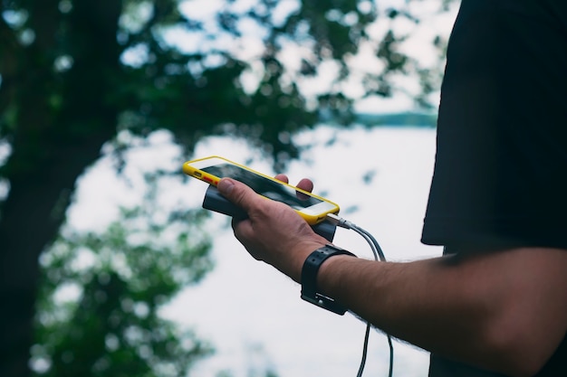 Le gars tient un chargeur portable avec un smartphone à la main. Homme sur fond de nature avec verdure et lac.