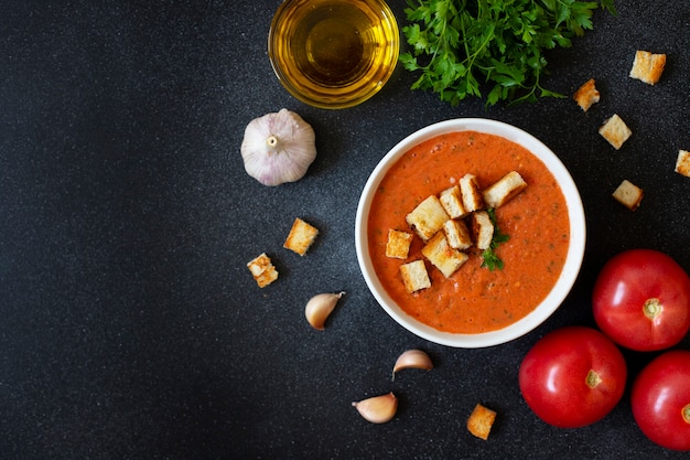 Gaspacho traditionnel de soupe froide aux tomates d'été servi dans un bol blanc. Tomates, ail, basilic, persil, huile d'olive et croûtons. Cuisine méditerranéenne, espagnole. Vue de dessus. Fond noir. Espace copie