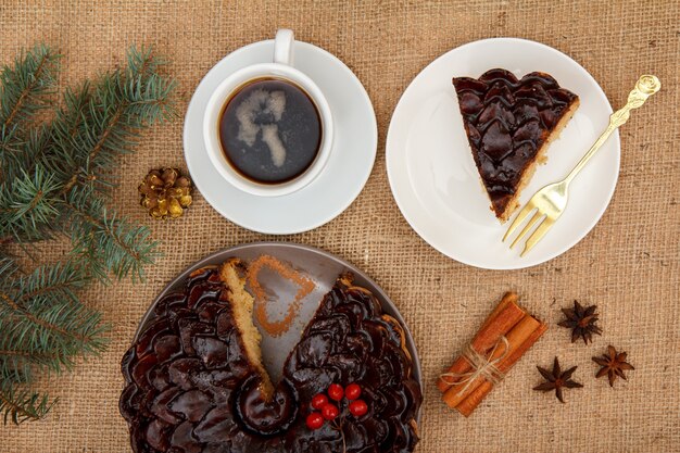 Gâteau au chocolat tranché décoré de bouquet de viorne, tasse de café, anis étoilé et cannelle sur table avec branche d'épinette et sac. Vue de dessus