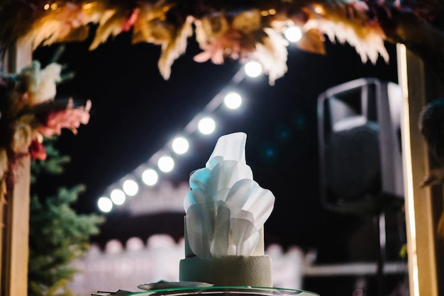 Gâteau aux fleurs blanches délicates la nuit avec lumière Cérémonie de mariage sous l'arche décorée de fleurs d'automne de l'extérieur dans la zone de banquet de l'arrière-cour Gâteau dans la zone de soirée magique Fête