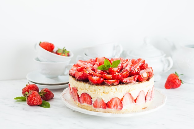 Gâteau aux fraises Fraisier sur l'assiette blanche et tasses pour l'heure du thé, baies et vaisselle en arrière-plan