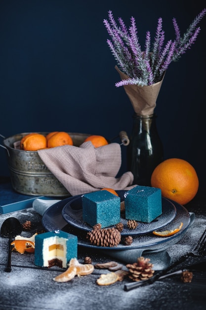 Gâteau Bleu Rempli D'orange Et De Mandarine En Forme De Cube. A Proximité Sur Une Table Noire Se Trouvent Des Ingrédients Oranges, Mandarines, Noix. Style Rustique