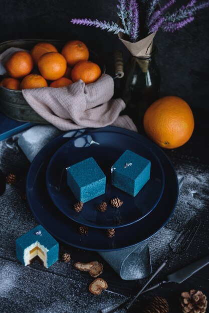 Gâteau bleu rempli d'orange et de mandarine en forme de cube. A proximité sur une table noire se trouvent des ingrédients oranges, mandarines, noix. Style rustique