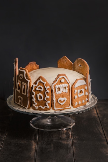 Gâteau fait maison avec des façades en pain d'épice de maisons de ville sur un support en verre et une table en bois sombre.