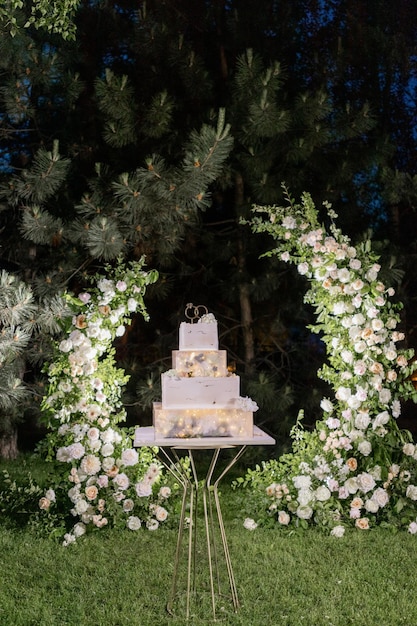 Photo un gâteau de mariage élégant étagé éclairé de lumières romantiques sur une toile de fond florale en plein air