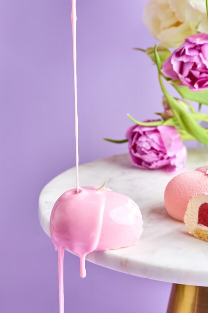 Un gâteau mousse est versé avec du glaçage sur un fond rose dans une composition avec des fleurs