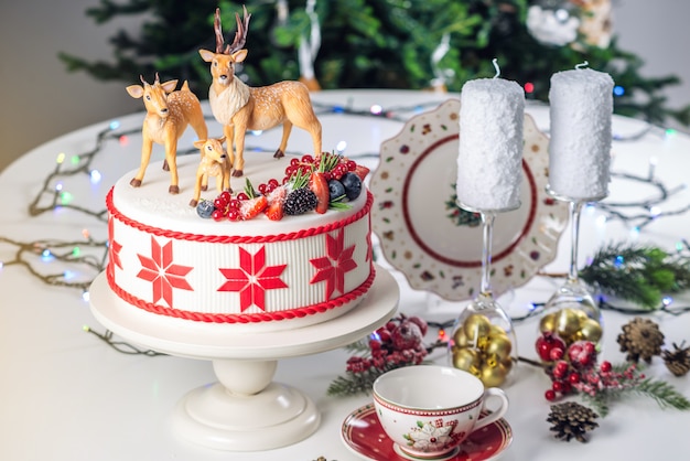 Gâteau de Noël blanc avec ornement rouge sur le dessus, décoré de cerfs et de baies fraîches sur une table de fête