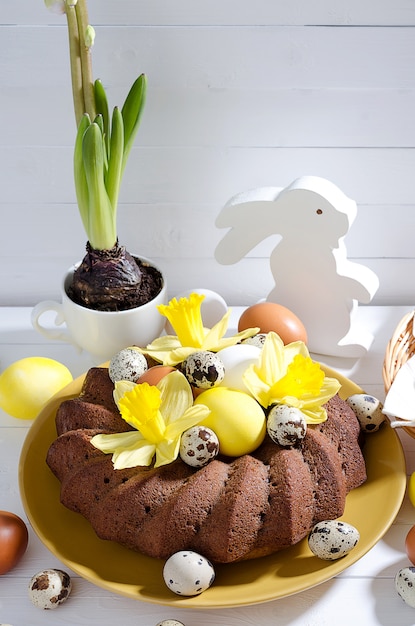 Gâteau de Pâques avec des œufs teints dans un nid, des jonquilles et une jacinthe dans une tasse sur un fond en bois rustique blanc