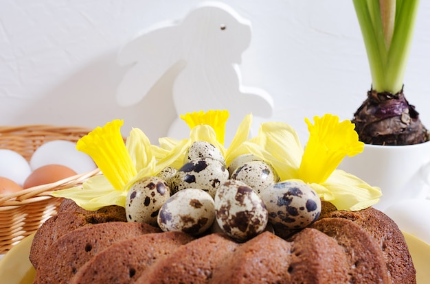 Photo gâteau de pâques avec des œufs teints dans un nid, des jonquilles et une jacinthe dans une tasse sur un fond en bois rustique blanc